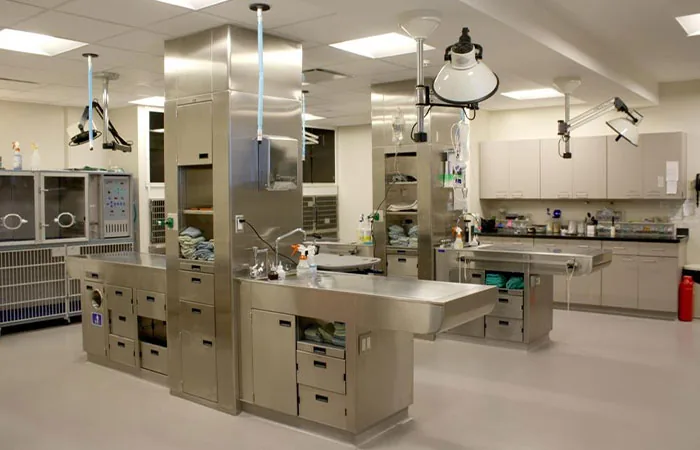 A modern, clean autopsy room with stainless steel tables, overhead lights, and built-in cabinets. Medical supplies and equipment are organized on countertops and shelves. The room is bright with neutral-colored walls and floors.