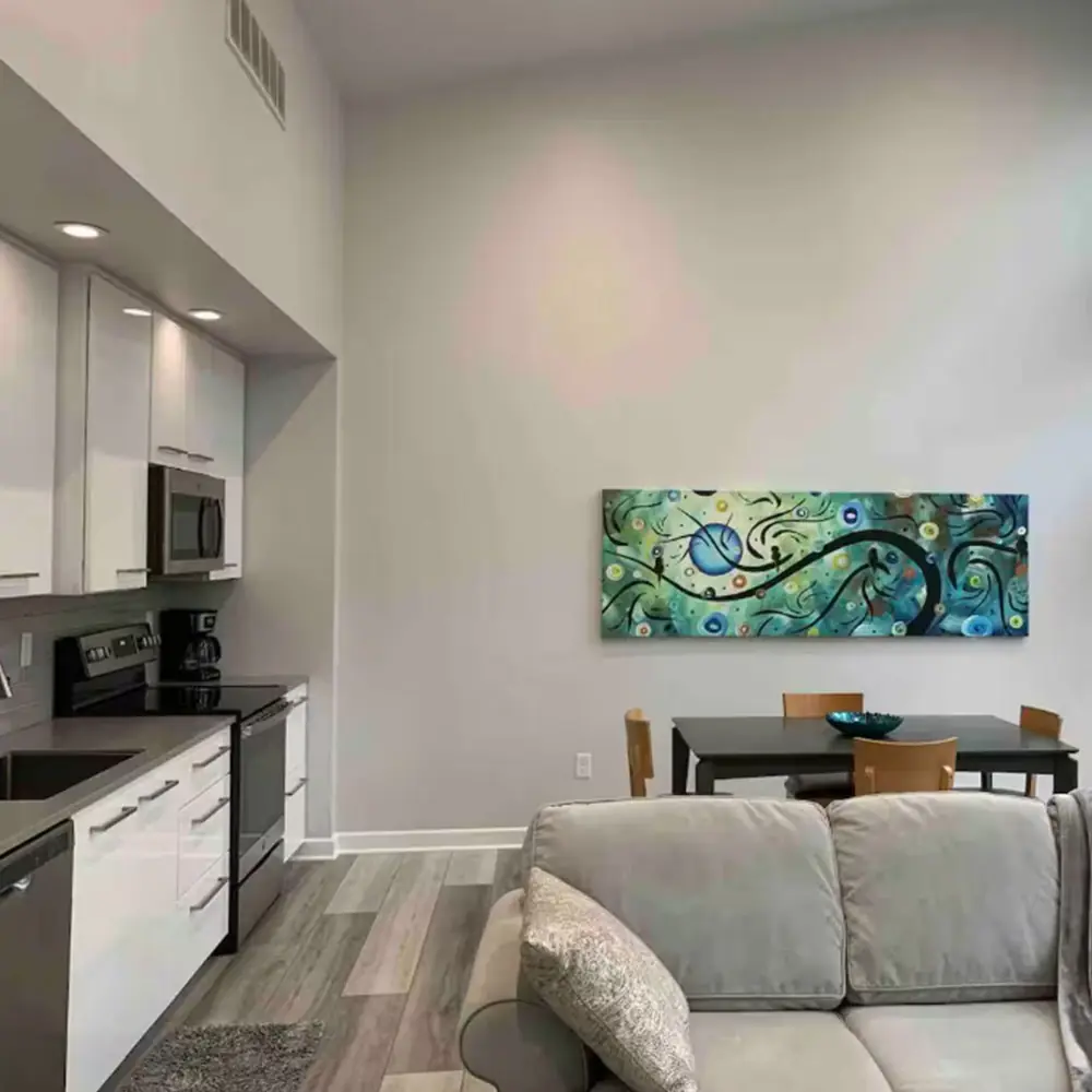 Modern apartment interior with a gray couch in the foreground, a dining table with four chairs and a blue bowl, vibrant abstract wall art, and a kitchen area with white cabinets and stainless steel appliances.