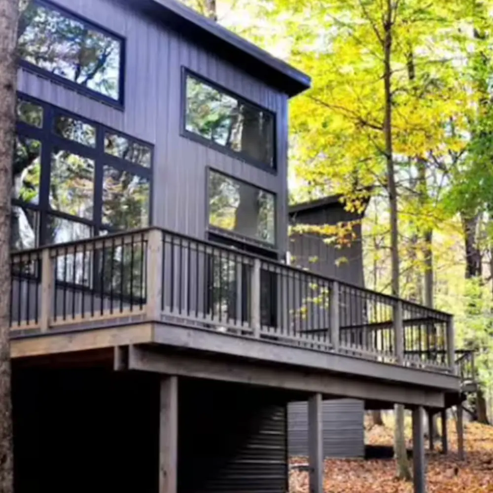 Modern grey cabin with large windows and a spacious wooden deck, situated among tall trees with green and yellow leaves in a forest setting during autumn.