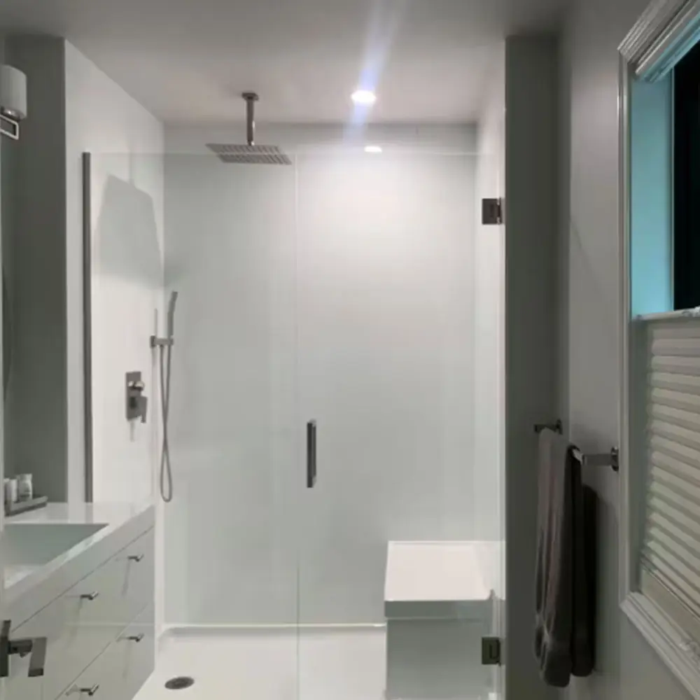 A modern, minimalistic bathroom with a glass-enclosed shower featuring a rain showerhead, a built-in white bench, white walls, a vanity with drawers on the left, and a towel hanging on the right wall.